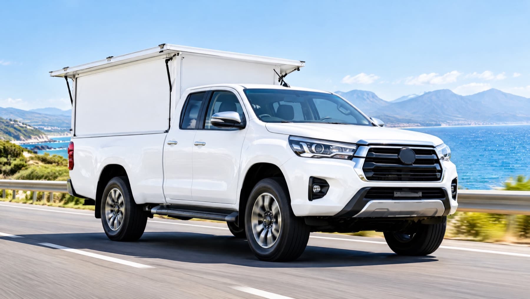A white double-cab bakkie with a white canopy driving along a sunny coastal road