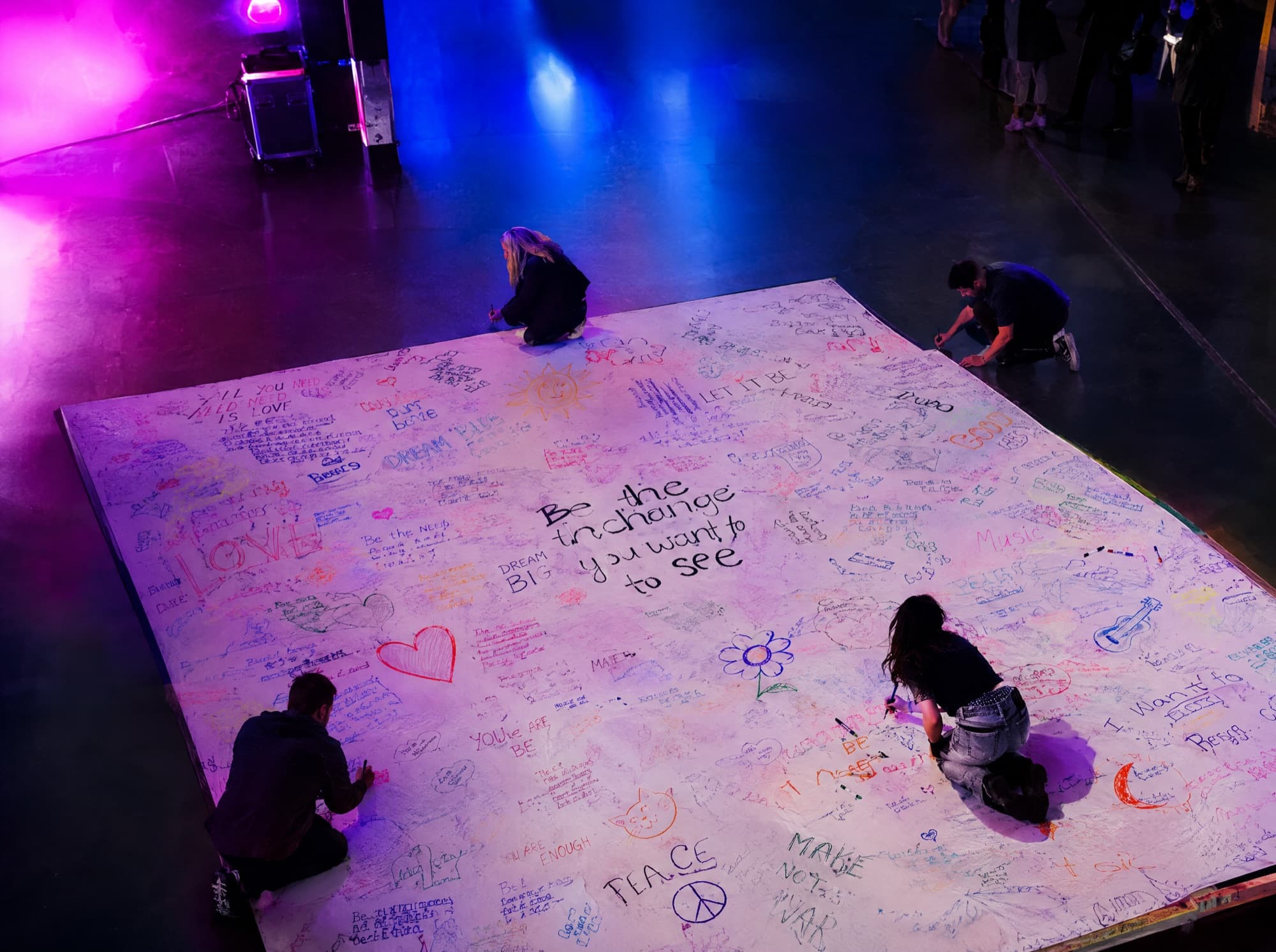 People kneeling on a giant white floor canvas, writing lyrics under coloured spotlights